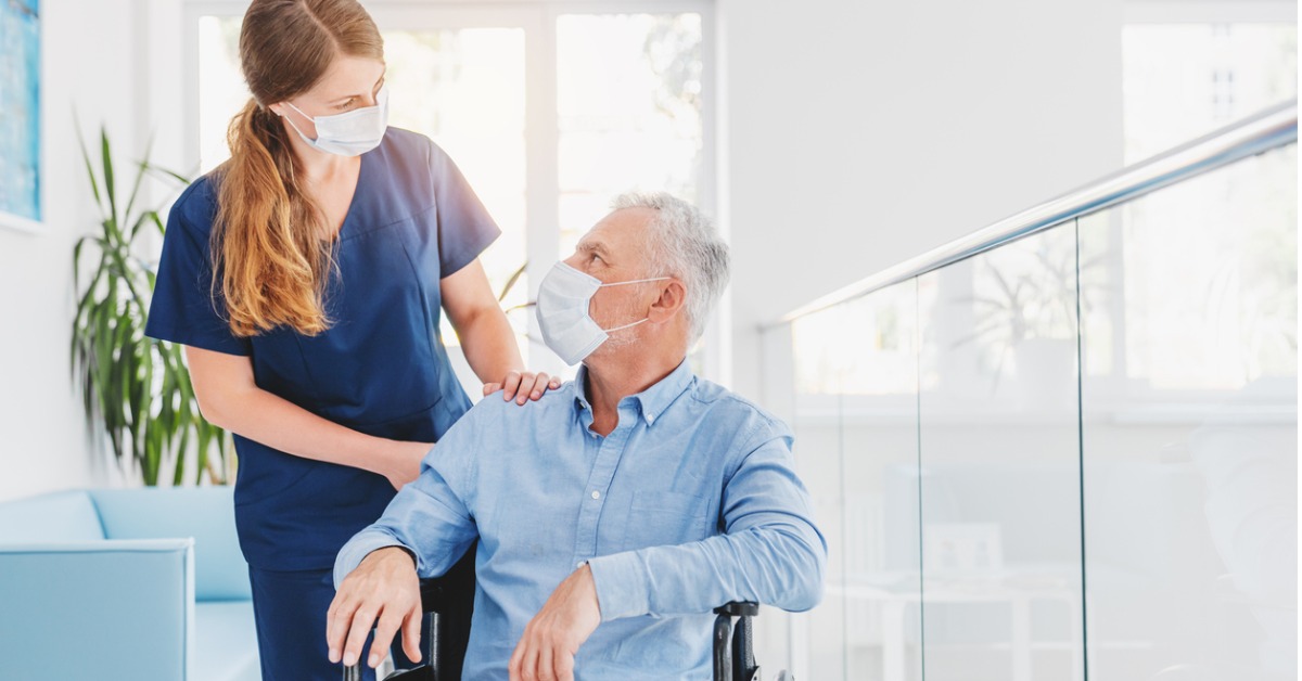 Young woman nurse talking to a patient in a wheelchair while wearing a face mask, in a hospital.