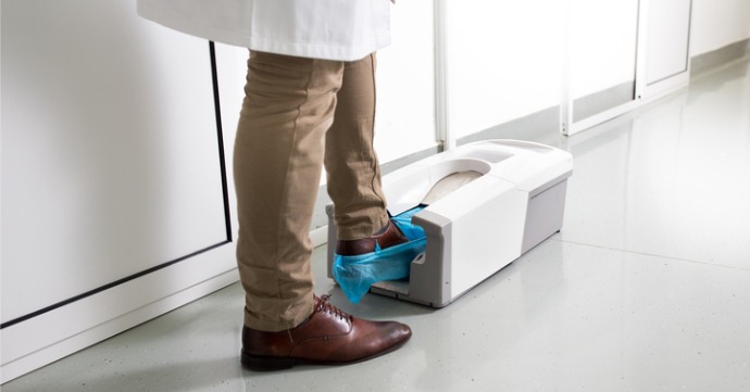 healthcare worker wearing a lab coat and using a shoe cover dispenser to put the shoe covers on